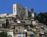 Vue d'un village perché dominé par les ruines imposantes du **château du marquis de Sade**, sous un ciel bleu éclatant.