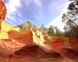 Carrières d'ocre aux couleurs vives, rappelant l'usine d'ocre Mathieu. Le ciel bleu et les arbres verts contrastent avec les roches rouges et jaunes.