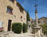 Village de pierre au soleil, le Crestet Vaucluse. Une croix orne la place pavée, entourée de maisons fleuries.