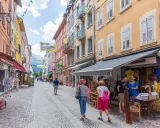 Rue commerçante animée à Aubenas-les-Alpes, avec des façades colorées, des drapeaux et des passants profitant de l'été.