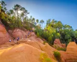 Des randonneurs explorent un sentier boisé aux ocres de Provence, baigné de soleil.