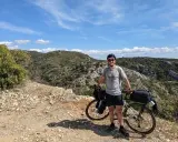 Cycliste souriant avec son vélo chargé, profitant d'une pause lors d'un tour du Luberon à vélo. Paysage rocailleux et collines verdoyantes sous un ciel bleu.