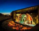 Spectacle son et lumière dans un théâtre antique. Le parvis orange illumine les gradins où le public est assis, tandis que la scène projette une fresque monumentale.