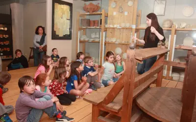 Visite d'un musée à Apt : des enfants écoutent attentivement une guide devant des étagères remplies d'objets anciens.