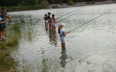 Des enfants pêchent dans le lac du Bartras, guidés par un adulte. L'eau reflète le ciel et la végétation.