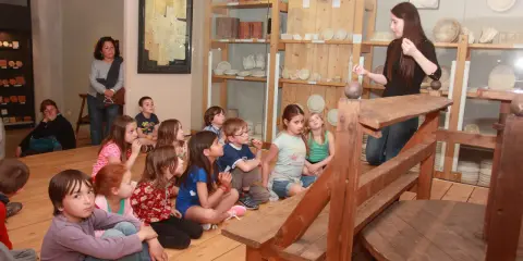Visite d'un musée à Apt : des enfants écoutent attentivement une guide devant des étagères remplies d'objets anciens.