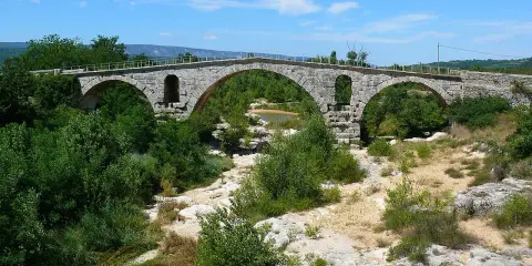 Vieux pont de pierre à arches sur une rivière asséchée, entouré de végétation luxuriante.
