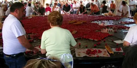 Des gens préparent une immense tarte aux fraises pour la fête de la fraise. Des fraises fraîches et des morceaux de pain sont disposés sur une grande table.