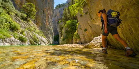 Randonneuse explorant la gorge du Toulourenc, traversant une rivière aux eaux cristallines bordée de falaises imposantes et de végétation luxuriante.