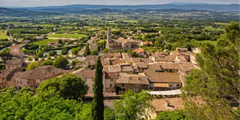 Vue aérienne du village de Bonnieux, avec ses toits de tuiles ocres, son église et la campagne environnante verdoyante. Le marché de Bonnieux est un lieu animé.