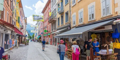 Rue commerçante animée à Aubenas-les-Alpes, avec des façades colorées, des drapeaux et des passants profitant de l'été.