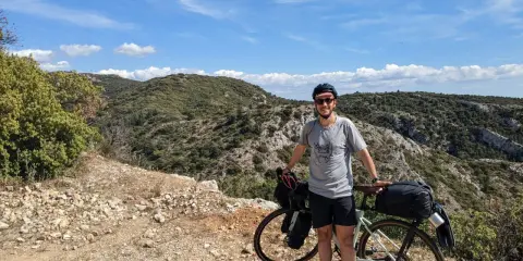 Cycliste souriant avec son vélo chargé, profitant d'une pause lors d'un tour du Luberon à vélo. Paysage rocailleux et collines verdoyantes sous un ciel bleu.