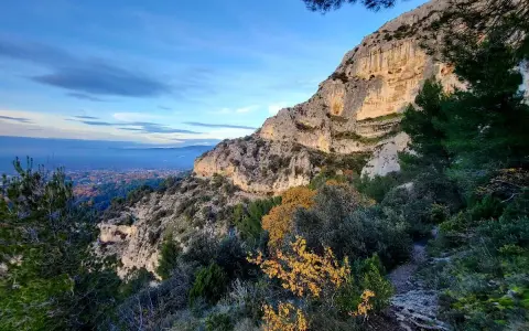 Vue panoramique des gorges de Badarel, avec un sentier rocailleux serpentant à travers la végétation méditerranéenne et une falaise imposante baignée de soleil.