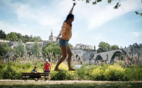Amusez-vous à Avignon ce week-end ! Une jeune femme saute devant le Palais des Papes et le Pont d'Avignon.