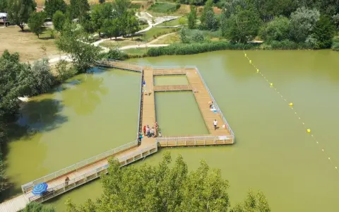 Ponton en bois sur un lac verdâtre, une **base de loisirs** animée avec des baigneurs profitant du soleil.