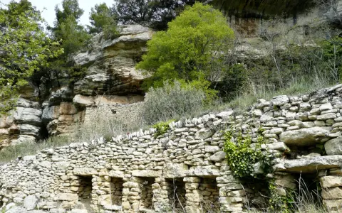 Vestiges d'une ancienne ville de murs en pierre, intégrée à une falaise rocheuse, avec végétation luxuriante.
