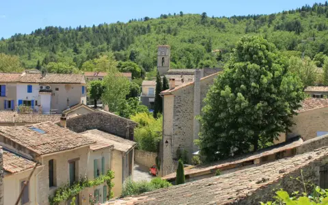 Vue d'un village provençal avec ses toits de tuiles, un clocher et une colline boisée. On imagine le marché de Cereste animé sous le soleil.