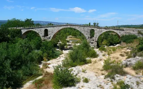 Vieux pont de pierre à arches sur une rivière asséchée, entouré de végétation luxuriante.