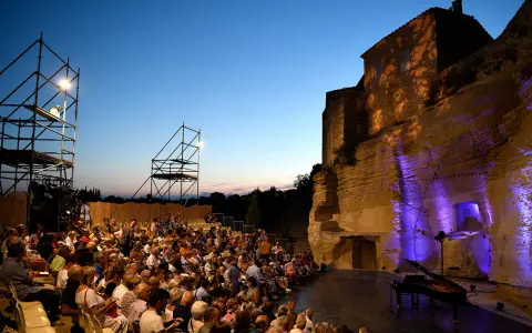 Public venu pour le festival de Gordes, une foule assise attend un concert au crépuscule, avec un piano sur scène et des structures métalliques.