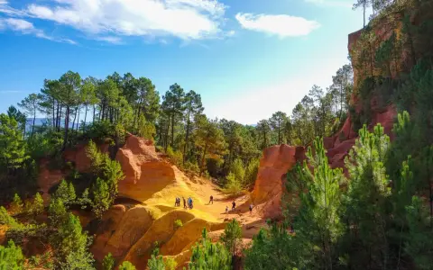 Des randonneurs explorent le sentier des ocres, un paysage spectaculaire aux falaises rouges et jaunes, bordé de pins verts sous un ciel bleu.