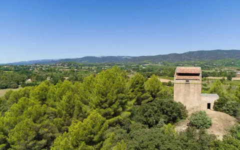 Vue panoramique sur la campagne provençale avec le moulin de Puyvert, entouré de pins et de verdure, sous un ciel bleu éclatant.