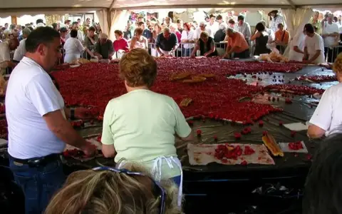 Des gens préparent une immense tarte aux fraises pour la fête de la fraise. Des fraises fraîches et des morceaux de pain sont disposés sur une grande table.