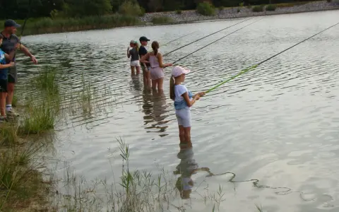 Des enfants pêchent dans le lac du Bartras, guidés par un adulte. L'eau reflète le ciel et la végétation.