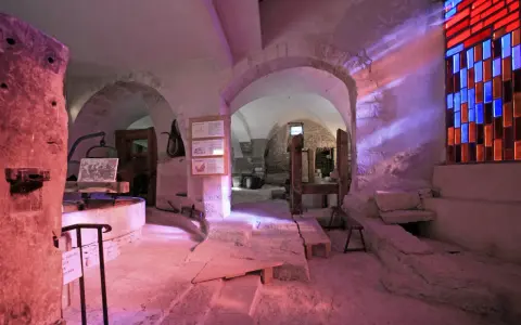 Intérieur d'un musée du verre et du vitrail, avec des arches en pierre, des outils anciens et un vitrail coloré projetant des rayons lumineux.