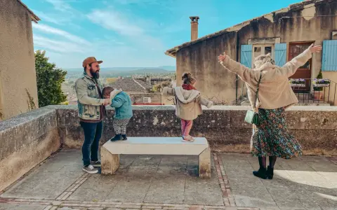 Famille profitant d'une vue panoramique depuis un balcon à Châteauneuf de Gadagne. Le père tient un enfant, tandis que deux autres enfants jouent sur un banc.