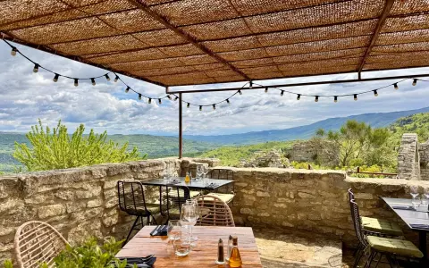 Terrasse d'un restaurant avec vue panoramique sur les collines. Tables dressées sous un auvent de chaume, ambiance estivale. Un jardin sur le toit Saignon promet un repas inoubliable.