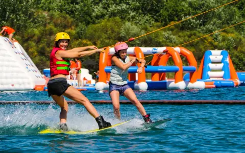 Deux femmes font du wakeboard dans un wam park orange et bleu, avec des structures gonflables en arrière-plan.