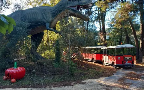 Un train rouge traverse le parc Grimmland, passant près d'un dinosaure géant et de citrouilles décoratives.