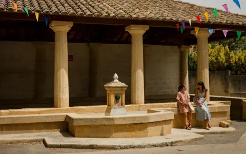 Deux femmes discutent près d'une fontaine sous un auvent orné de fanions colorés, ambiance de marché à Rochefort-du-Gard.