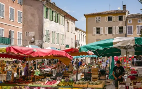 Le marché le Thor animé, avec des étals colorés de fruits et légumes sous des parasols. Des passants flânent entre les stands, profitant du soleil provençal.