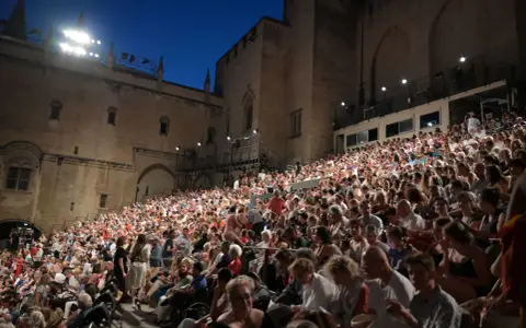 Foule nombreuse assise sur des gradins, devant un palais, pour la Fête de la Musique à Avignon.