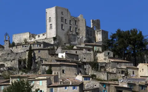 Vue d'un village perché dominé par les ruines imposantes du **château du marquis de Sade**, sous un ciel bleu éclatant.