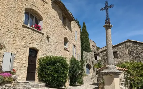Village de pierre au soleil, le Crestet Vaucluse. Une croix orne la place pavée, entourée de maisons fleuries.