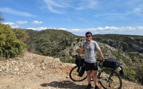 Cycliste souriant avec son vélo chargé, profitant d'une pause lors d'un tour du Luberon à vélo. Paysage rocailleux et collines verdoyantes sous un ciel bleu.