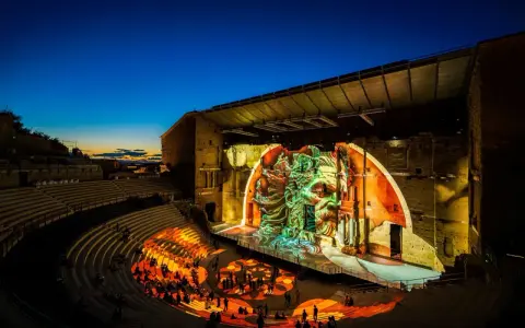 Spectacle son et lumière dans un théâtre antique. Le parvis orange illumine les gradins où le public est assis, tandis que la scène projette une fresque monumentale.