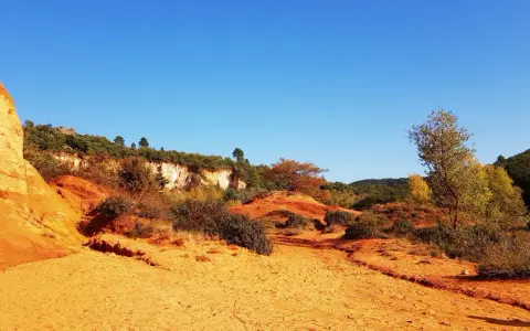 Paysage ocre et rougeoyant du Colorado provençal, avec des falaises, des arbustes et un ciel bleu intense.
