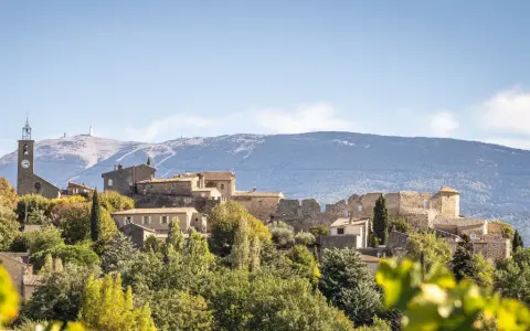 Vue d'un faucon village perché sur une colline, avec une église et des ruines médiévales. Le Mont Ventoux domine l'horizon.