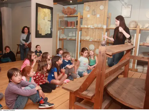 Visite d'un musée à Apt : des enfants écoutent attentivement une guide devant des étagères remplies d'objets anciens.