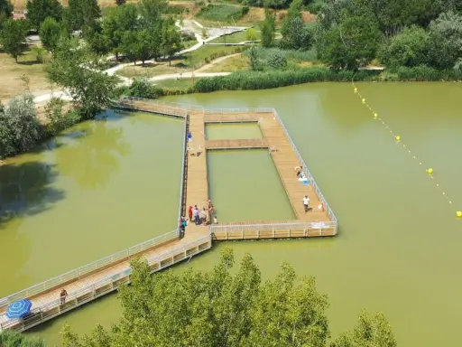 Ponton en bois sur un lac verdâtre, une **base de loisirs** animée avec des baigneurs profitant du soleil.