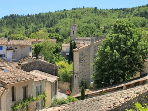 Vue d'un village provençal avec ses toits de tuiles, un clocher et une colline boisée. On imagine le marché de Cereste animé sous le soleil.