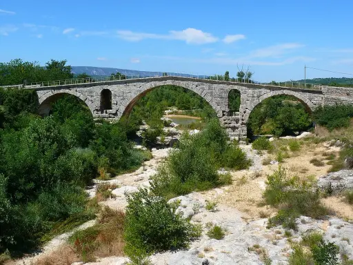 Vieux pont de pierre à arches sur une rivière asséchée, entouré de végétation luxuriante.