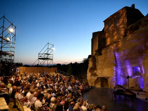 Public venu pour le festival de Gordes, une foule assise attend un concert au crépuscule, avec un piano sur scène et des structures métalliques.