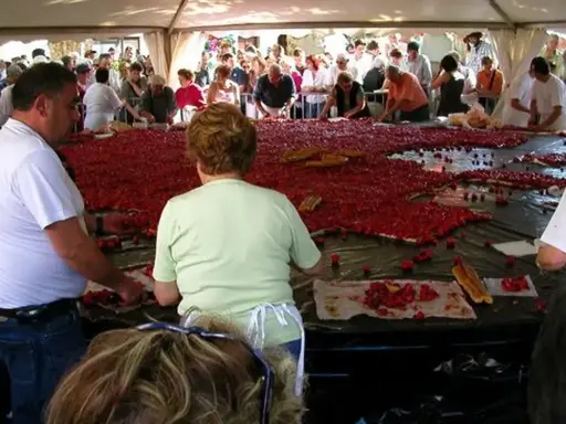 Des gens préparent une immense tarte aux fraises pour la fête de la fraise. Des fraises fraîches et des morceaux de pain sont disposés sur une grande table.