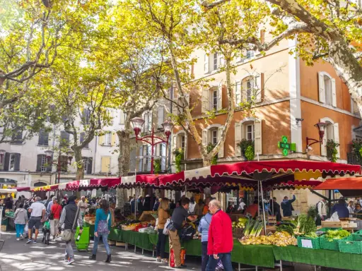 Ambiance animée au marché de Manosque, sous les platanes aux feuilles d'automne. Étals colorés de fruits et légumes frais, passants flânant.