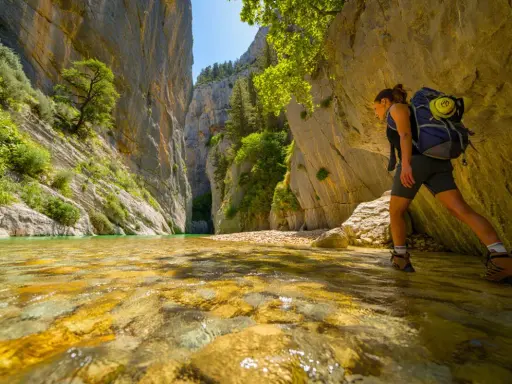 Randonneuse explorant la gorge du Toulourenc, traversant une rivière aux eaux cristallines bordée de falaises imposantes et de végétation luxuriante.