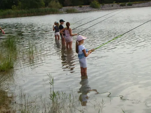 Des enfants pêchent dans le lac du Bartras, guidés par un adulte. L'eau reflète le ciel et la végétation.